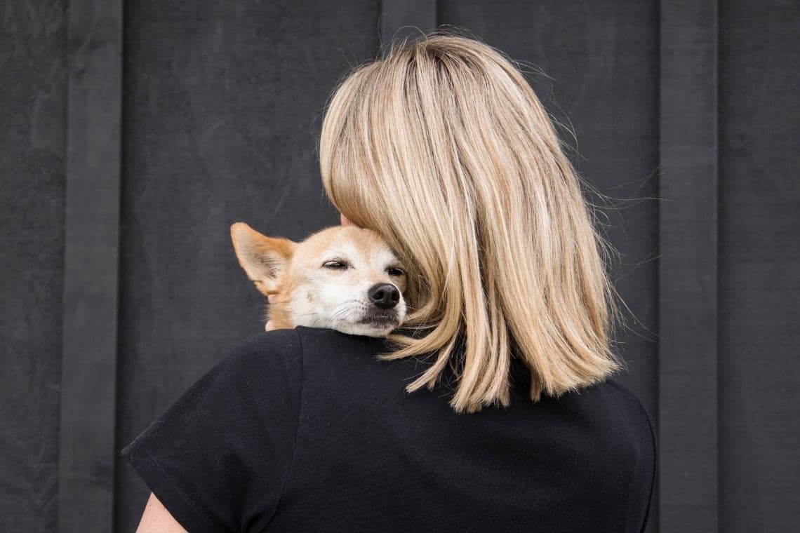 A blonde woman holds a small, tan dog with closed eyes on her shoulder.